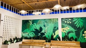 Een kamer met een hoge plafond, houten balken en een groene, bladerige muurschildering. Er staan houten banken en een witte lattenwand.
