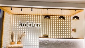 Een receptiegedeelte met een houten receptiebalie, een muur met de tekst 'HOLA BAY' in grote letters, en twee potplanten.