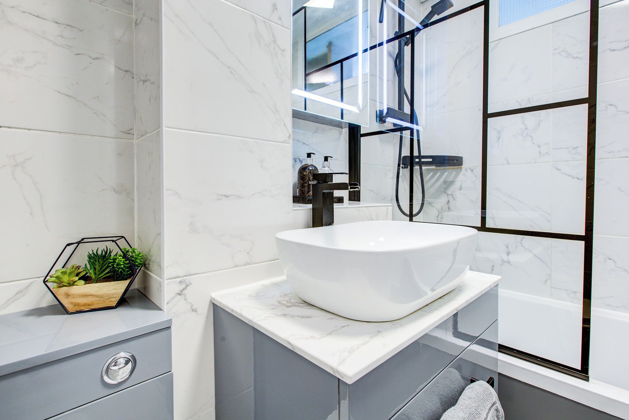 Un baño moderno con lavamanos de mármol blanco, una maceta hexagonal con plantas y un box de ducha de vidrio.