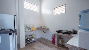 A compact kitchen with a refrigerator, a water dispenser, a gas stove, and a shelf with kitchen utensils. - SDK Homestay Uluwatu RedPartner (Bali)