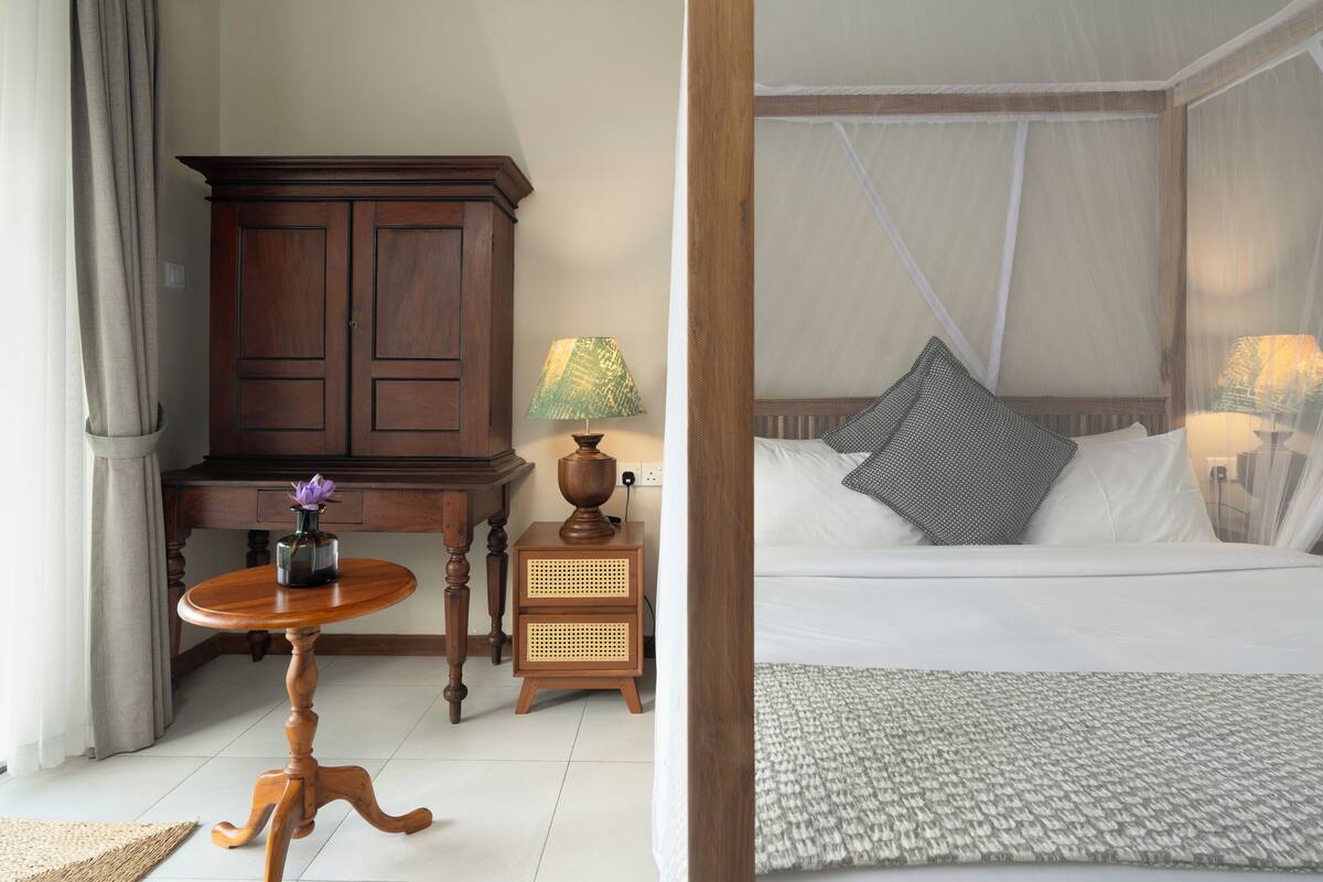 Une chambre d’hôtel avec un meuble en bois, une petite table ronde ornée d’un vase, une lampe et un lit avec une literie blanche et un baldaquin.