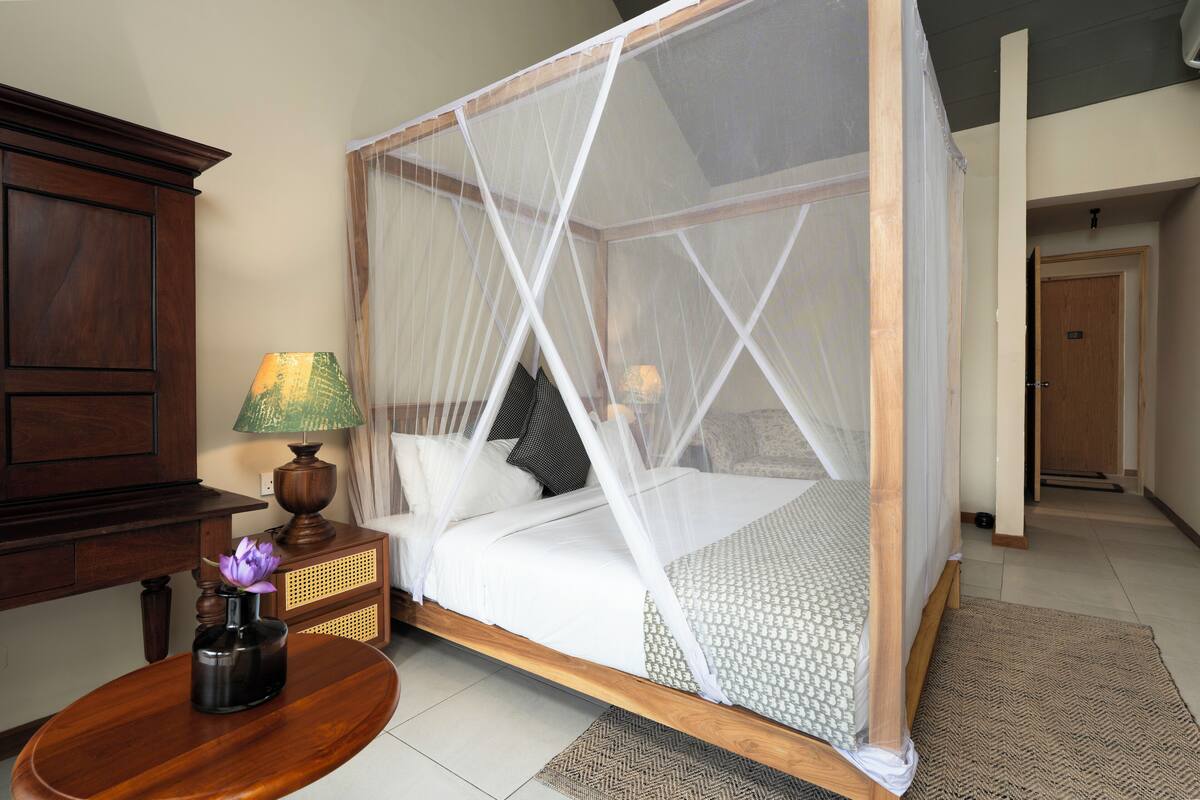 Un lit à baldaquin avec des rideaux blancs, une table de chevet en bois avec une lampe et un petit vase avec des fleurs.