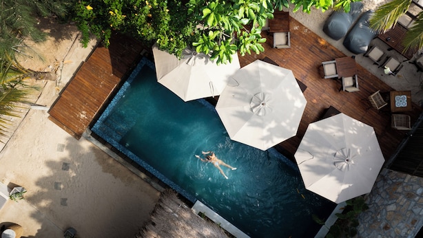 Une vue aérienne d’une piscine où une personne nage, entourée d’une terrasse en bois, de parasols blancs et de chaises longues.