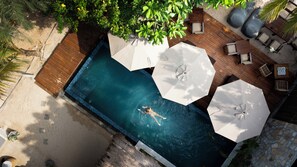Une vue aérienne d’une piscine où une personne nage, entourée d’une terrasse en bois, de parasols blancs et de chaises longues.