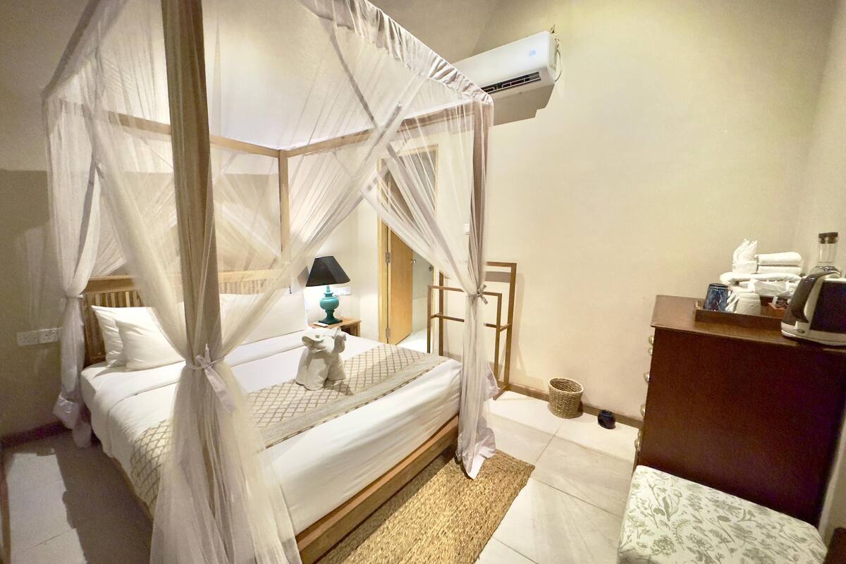 Une chambre à coucher avec un lit à baldaquin, une lampe de chevet, une petite statue et un bureau équipé d’un ordinateur et de produits de toilette.