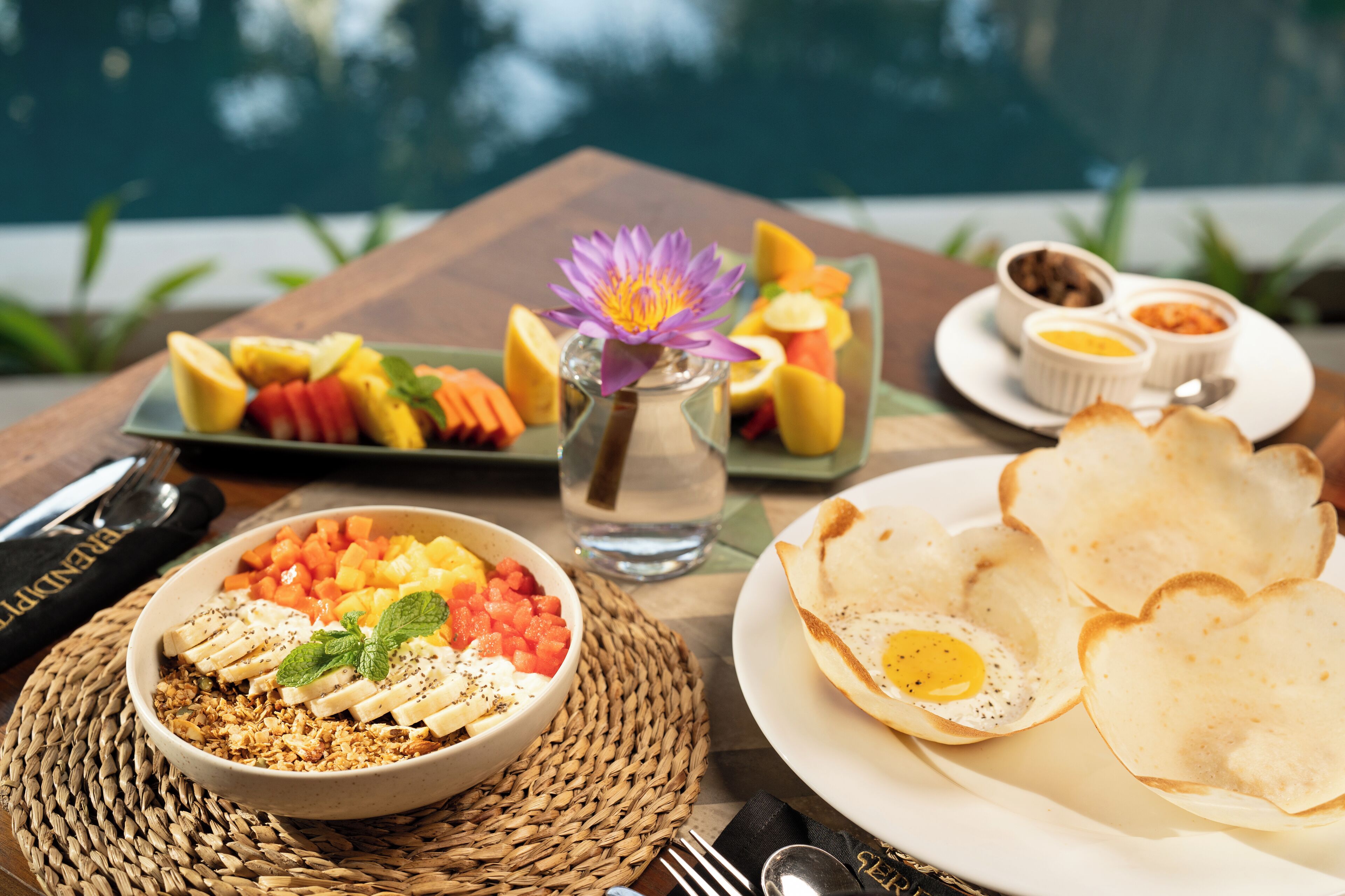 A breakfast setting with a bowl of granola, fruits, and a flower on a woven mat.