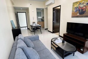 Un salon moderne avec un canapé, une table basse et un téléviseur à écran plat. On y trouve également un coin repas avec une table et des chaises, et une cuisine à l’arrière-plan.