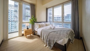 Une chambre à coucher avec un lit, une table de chevet, une lampe, une plante et de grandes fenêtres avec des rideaux.