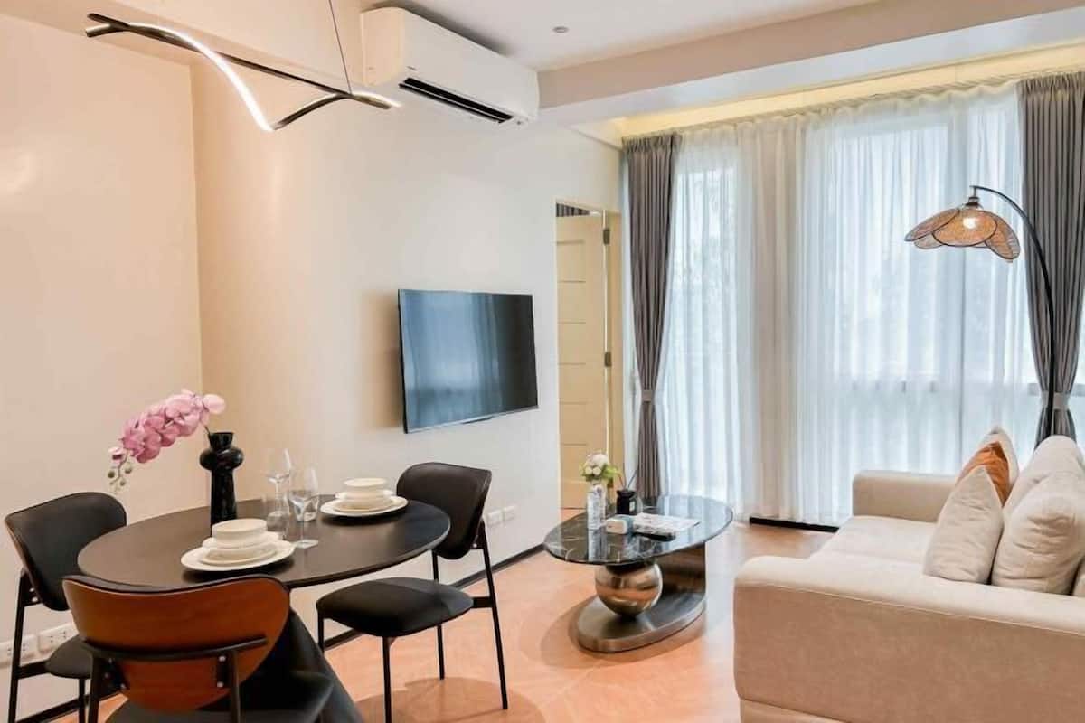 Ein modernes Wohnzimmer mit einer Couch, einem Esstisch mit Stühlen, einem Flachbildfernseher und einem großen Fenster mit Vorhängen.