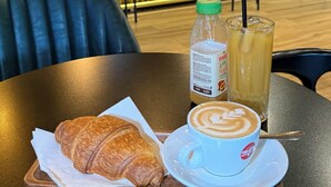 A café setting with a table set for a meal, including a croissant, a cup of coffee, and a bottle of apple cider. - ES Hotel (Kashar)