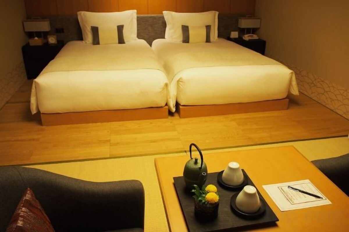A hotel room with two beds, a small table with a teapot, and a nightstand with a lamp.