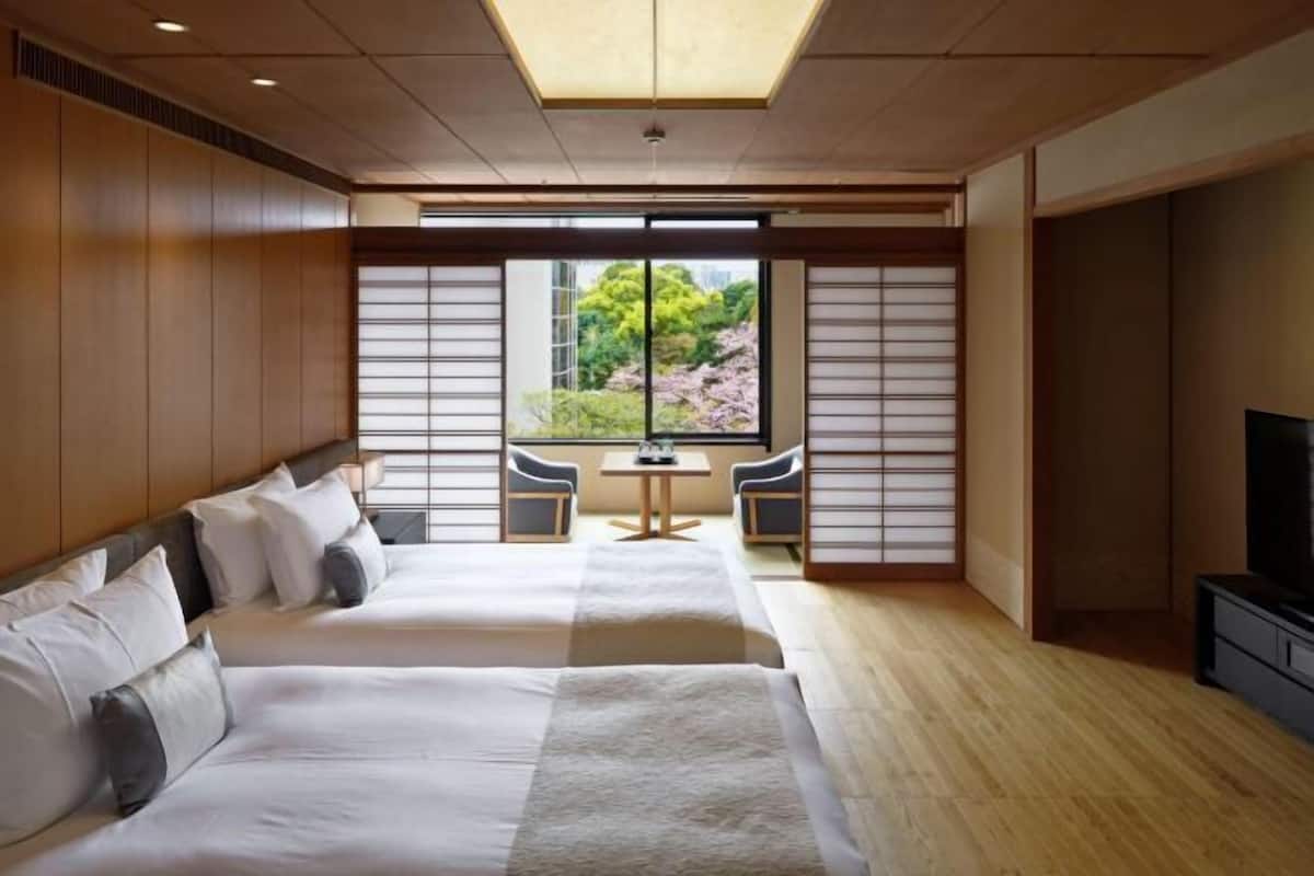A hotel room with two beds, a TV, a small table, and a view of greenery outside.