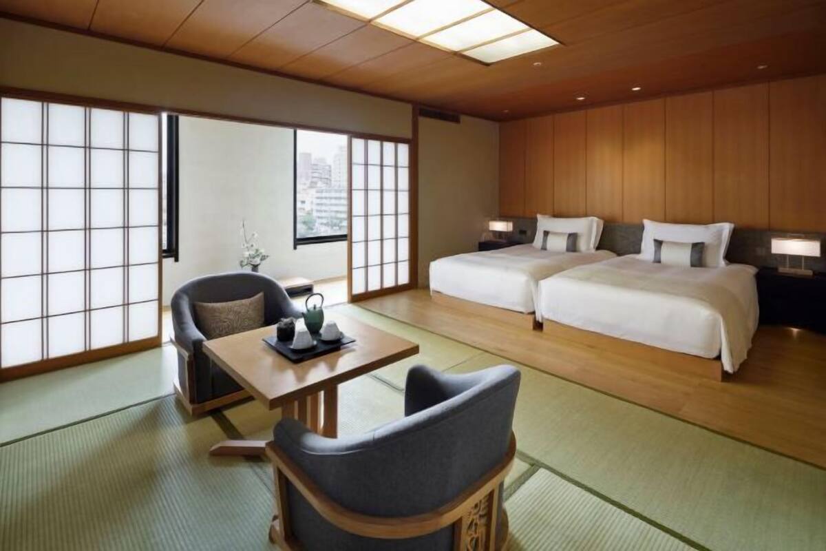 A traditional Japanese-style hotel room with tatami flooring, a low table with a tea set, two beds with pillows, and sliding shoji doors.