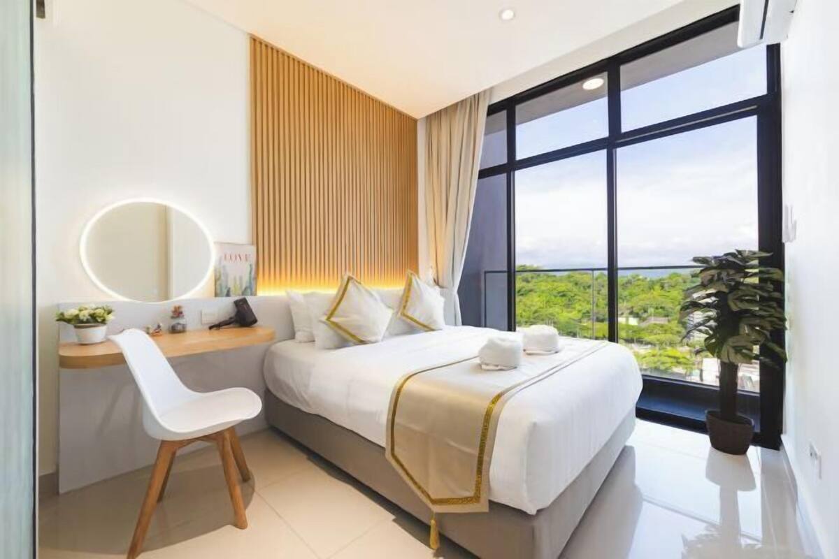 Ein modernes Hotelzimmer mit einem großen Bett, einem Schreibtisch mit Stuhl, einem runden Spiegel und Blick auf Grünflächen durch große Fenster.