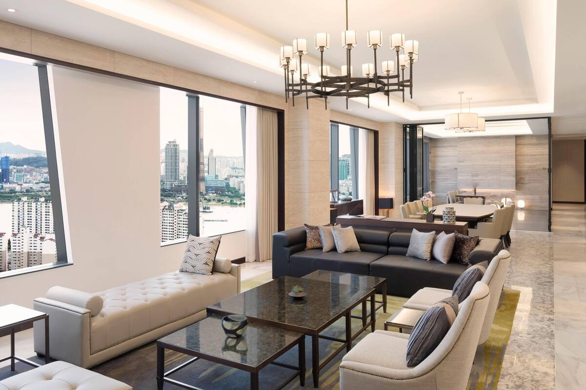 A modern hotel room with a city view, a large sofa, a dining area, and a chandelier.
