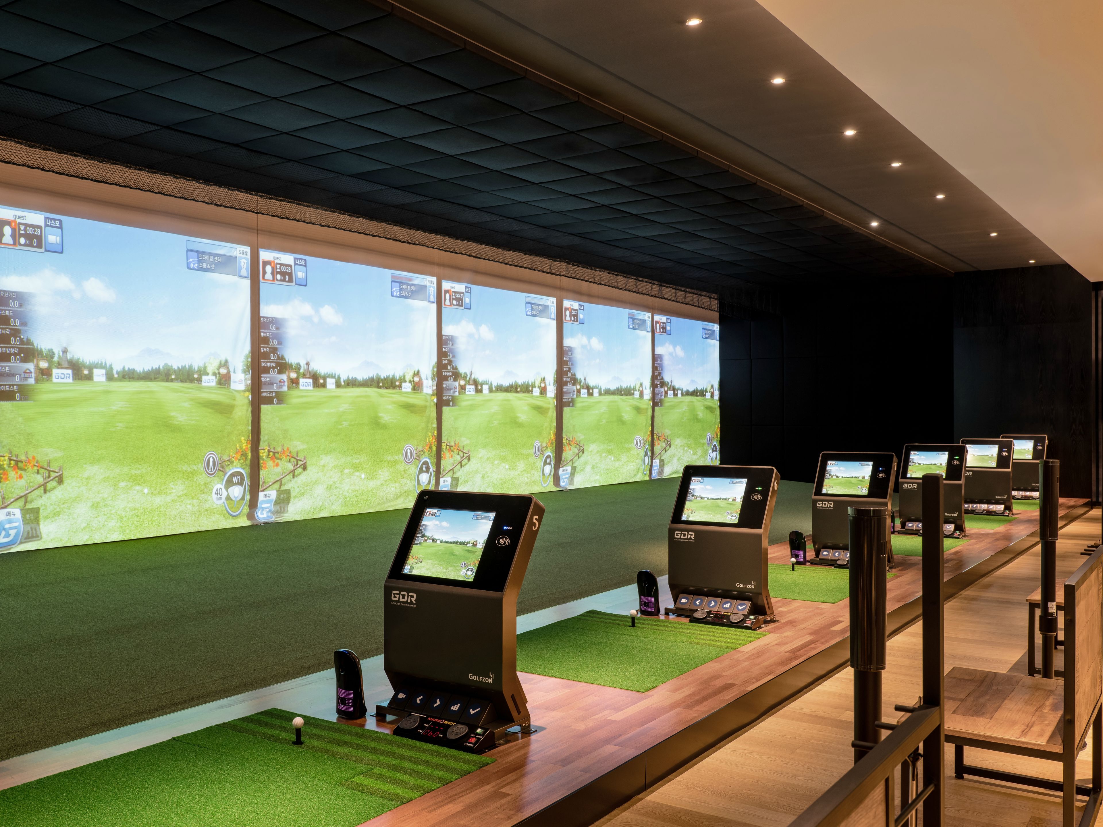 A golf simulation room with multiple screens displaying golf courses, a wooden floor, and a ceiling with a grid pattern.