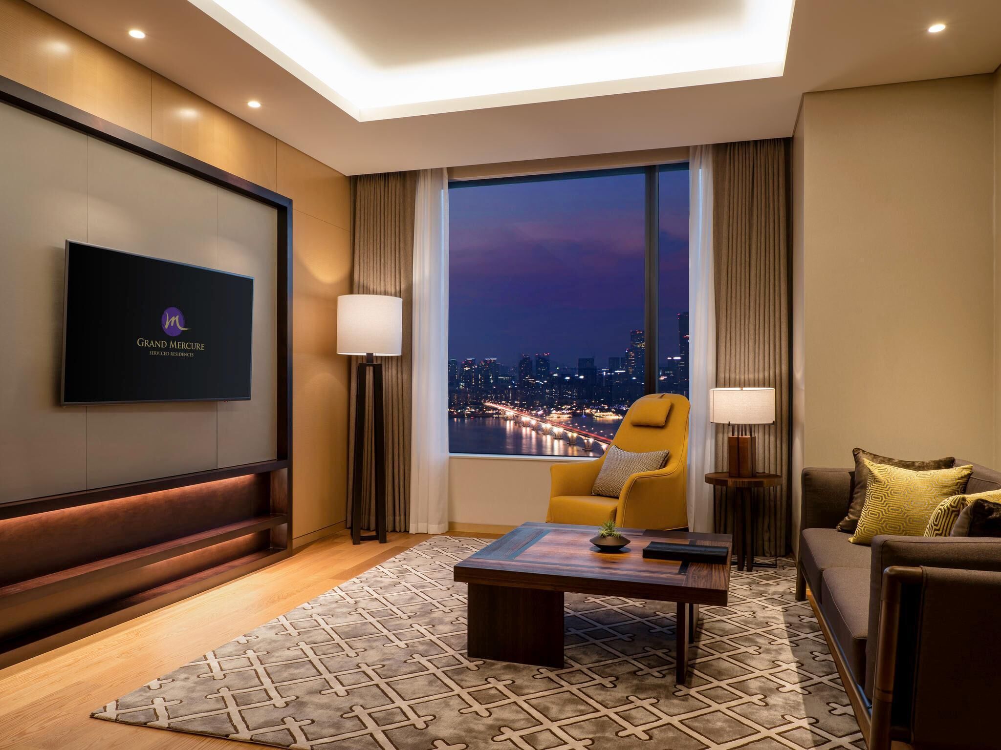 Une chambre d’hôtel avec vue sur la ville, un téléviseur à écran plat, un canapé, un fauteuil, une table basse et un tapis à motifs.