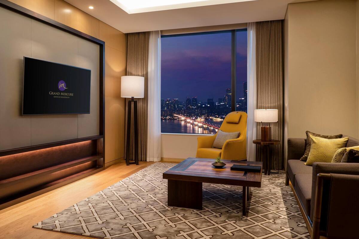 A hotel room with a city view, a flat-screen TV, a sofa, a chair, a coffee table, and a patterned rug.