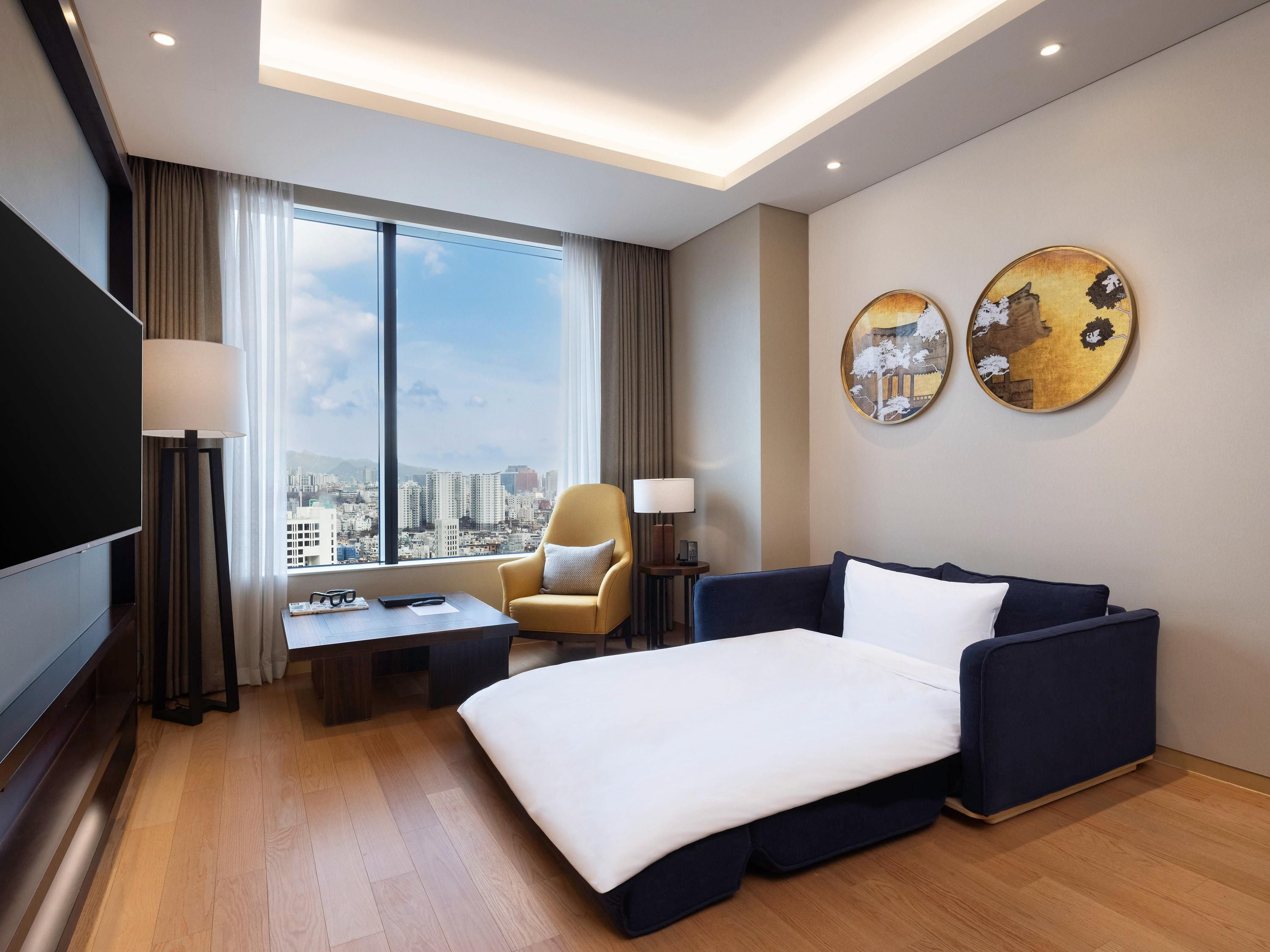 A hotel room with a bed, a sofa, a desk, a chair, a TV, and a large window with a city view.
