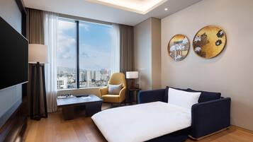 A hotel room with a bed, a sofa, a desk, a chair, a TV, and a large window with a city view.