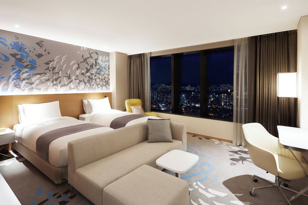 A hotel room with two beds, a sofa, a small table, a chair, and a city view.