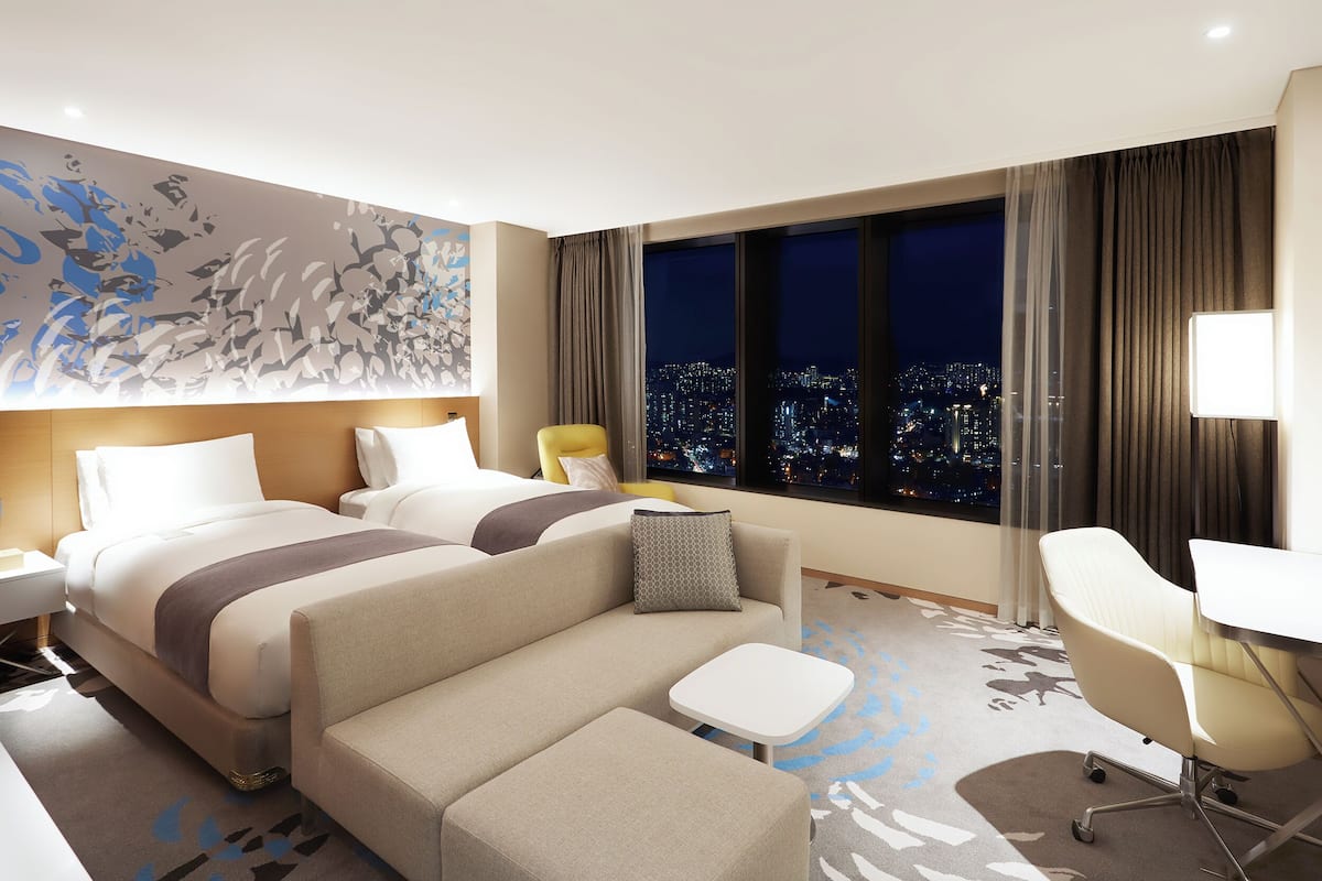 Une chambre d’hôtel avec deux lits, un canapé, une petite table, une chaise et une vue sur la ville.