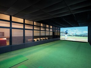 Quarto com uma tela grande exibindo um jogo de beisebol, piso coberto por carpete verde e uma área de estar com mesas e cadeiras.