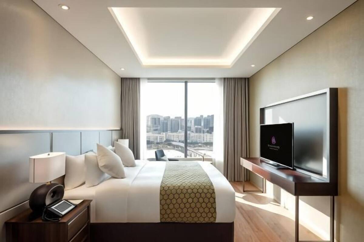 A modern hotel room with a large bed, a desk with a laptop, a television, and a city view.