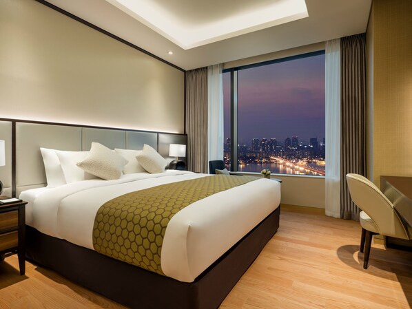 A modern hotel room with a large bed, a desk, and a view of a cityscape at dusk.