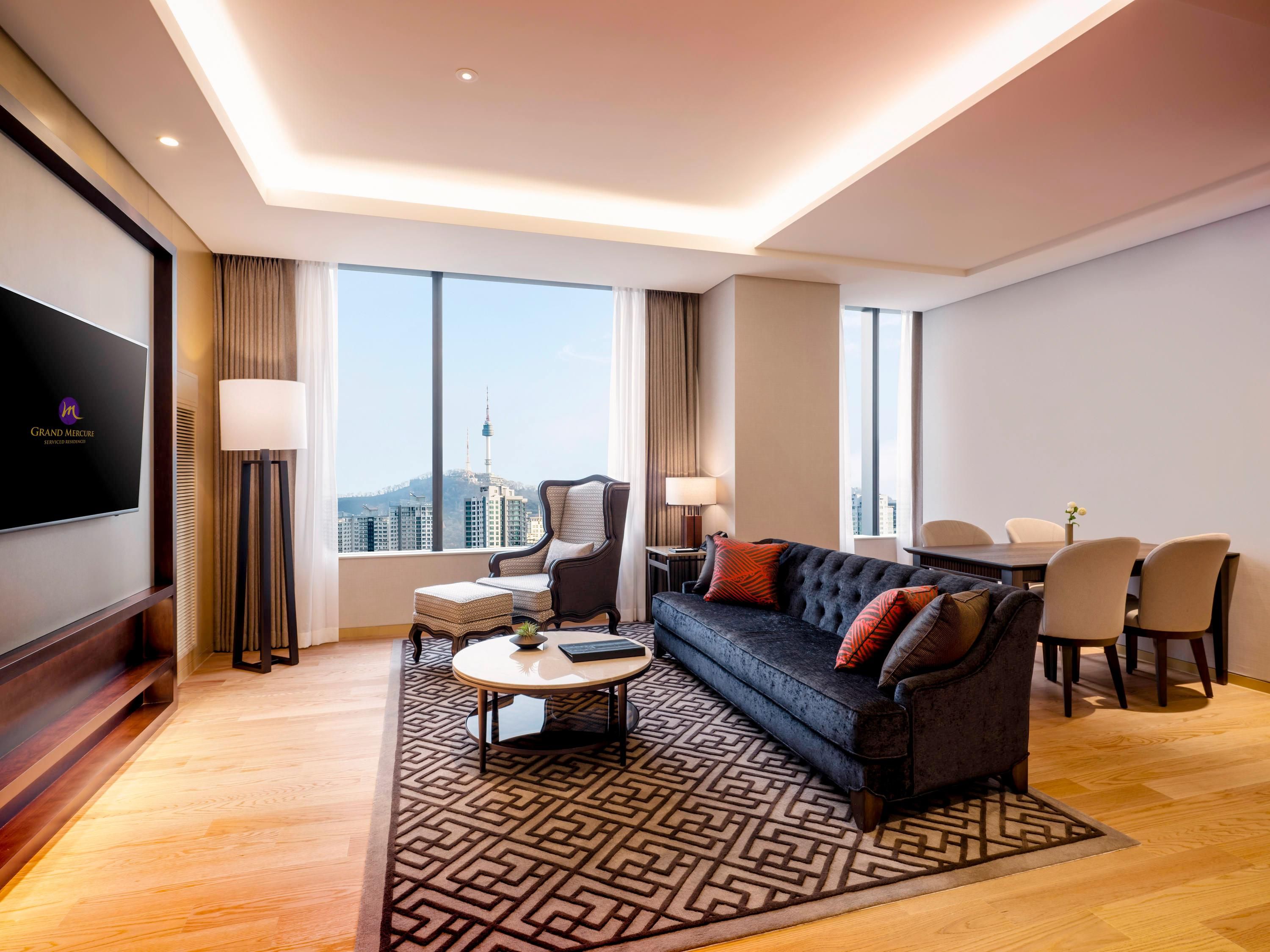 Una habitación de hotel moderna con sofá, mesa de comedor, sillas, televisor y vistas a la ciudad.