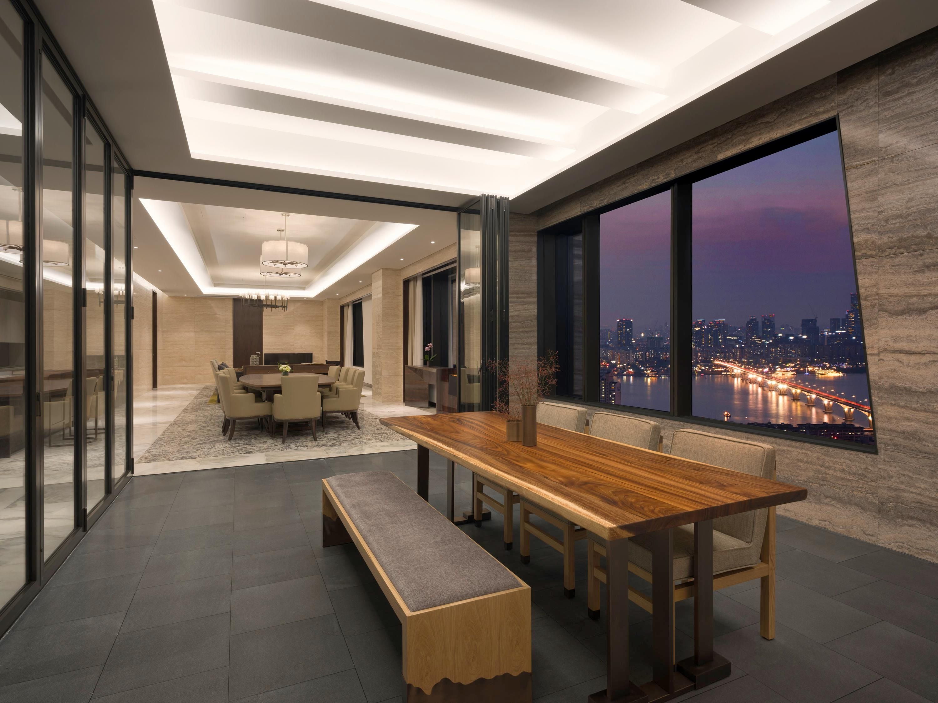 A modern dining area with a long wooden table, benches, and a city view through large windows.