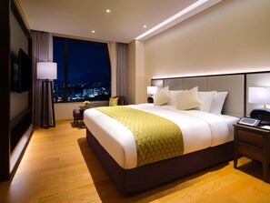 Quarto de hotel com uma cama grande, mesas de cabeceira, escrivaninha, cadeira, televisão e vista para a cidade.