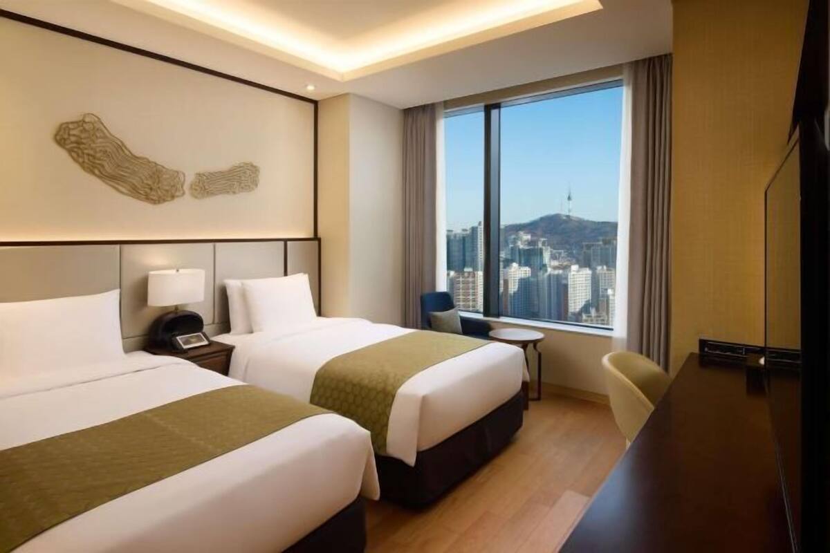 A hotel room with two beds, a desk, a chair, a small table, and a large window with a city view.
