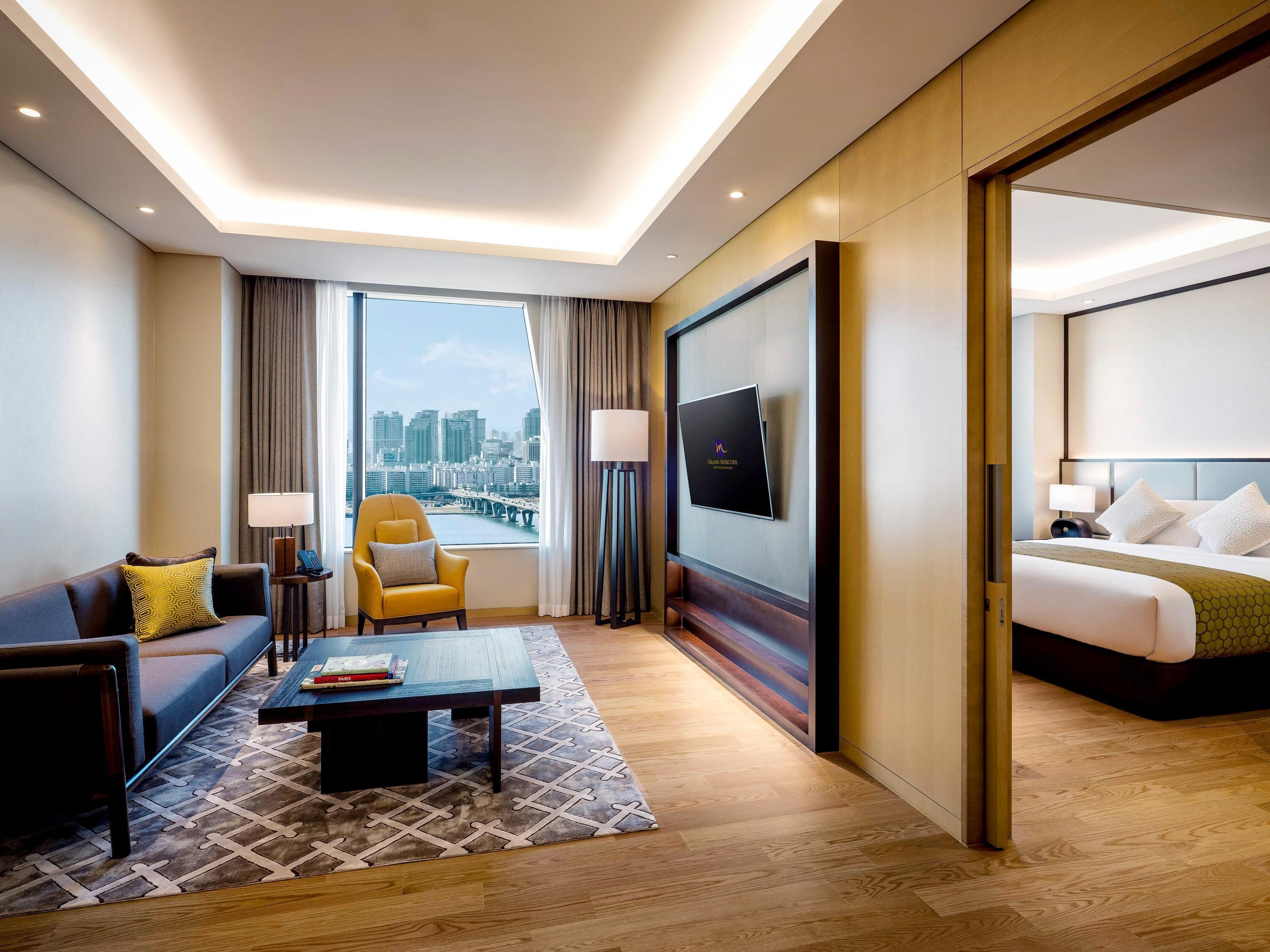 Une chambre d’hôtel comprenant un lit, un canapé, un fauteuil, une table basse, une télévision et une grande fenêtre donnant sur la ville.