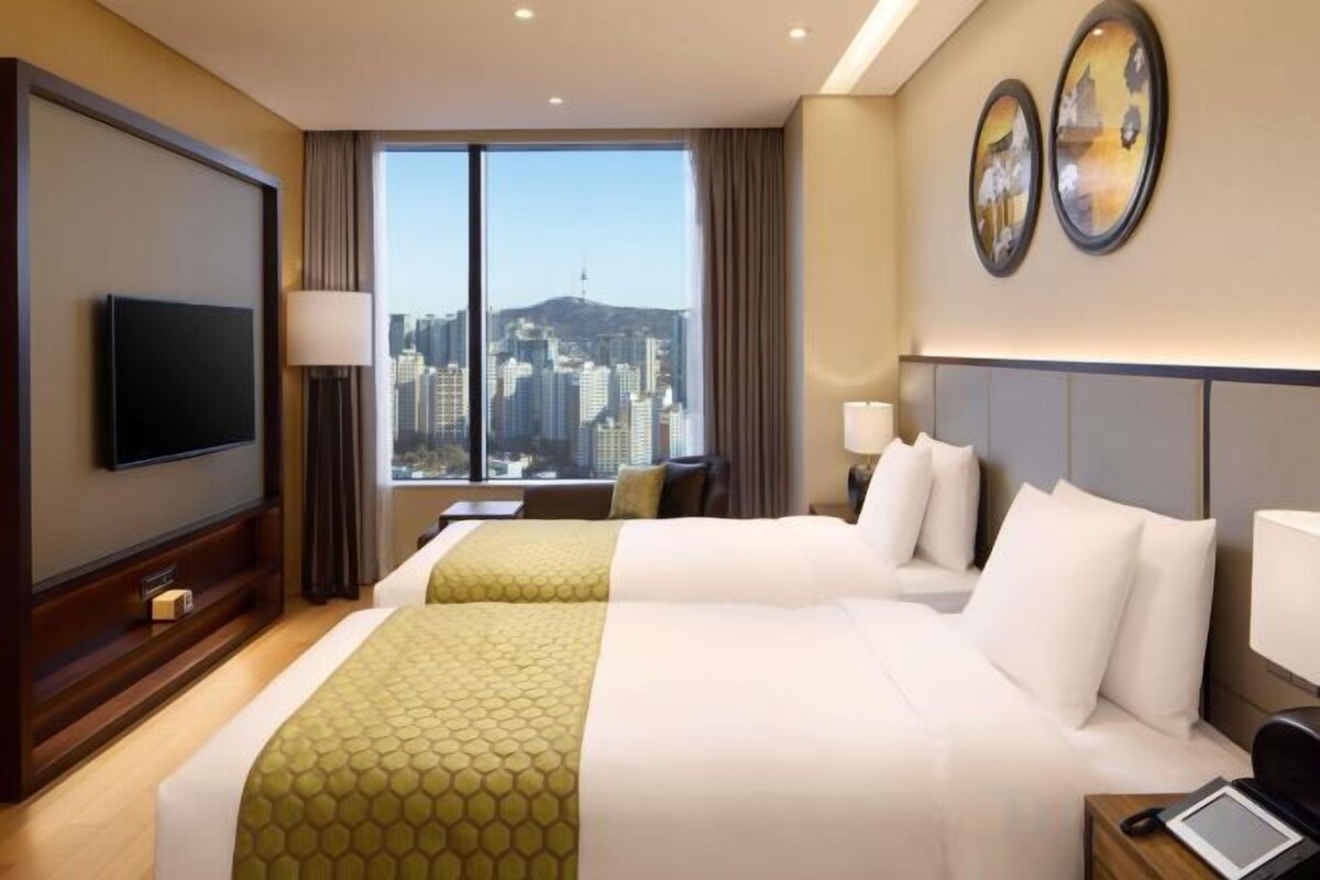 A hotel room with two beds, a TV, a desk, and a view of the city.