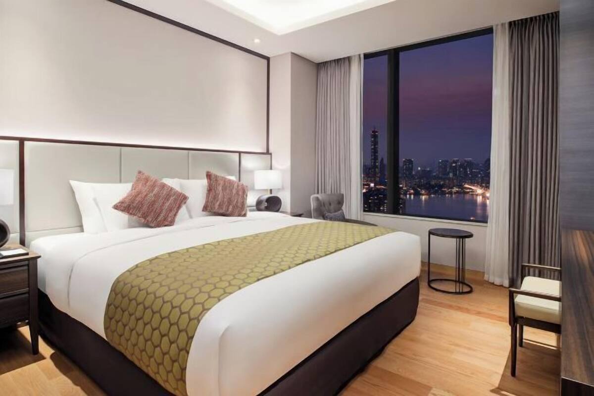 A modern hotel room with a large bed, a view of a cityscape at dusk, a small round table, and a chair.
