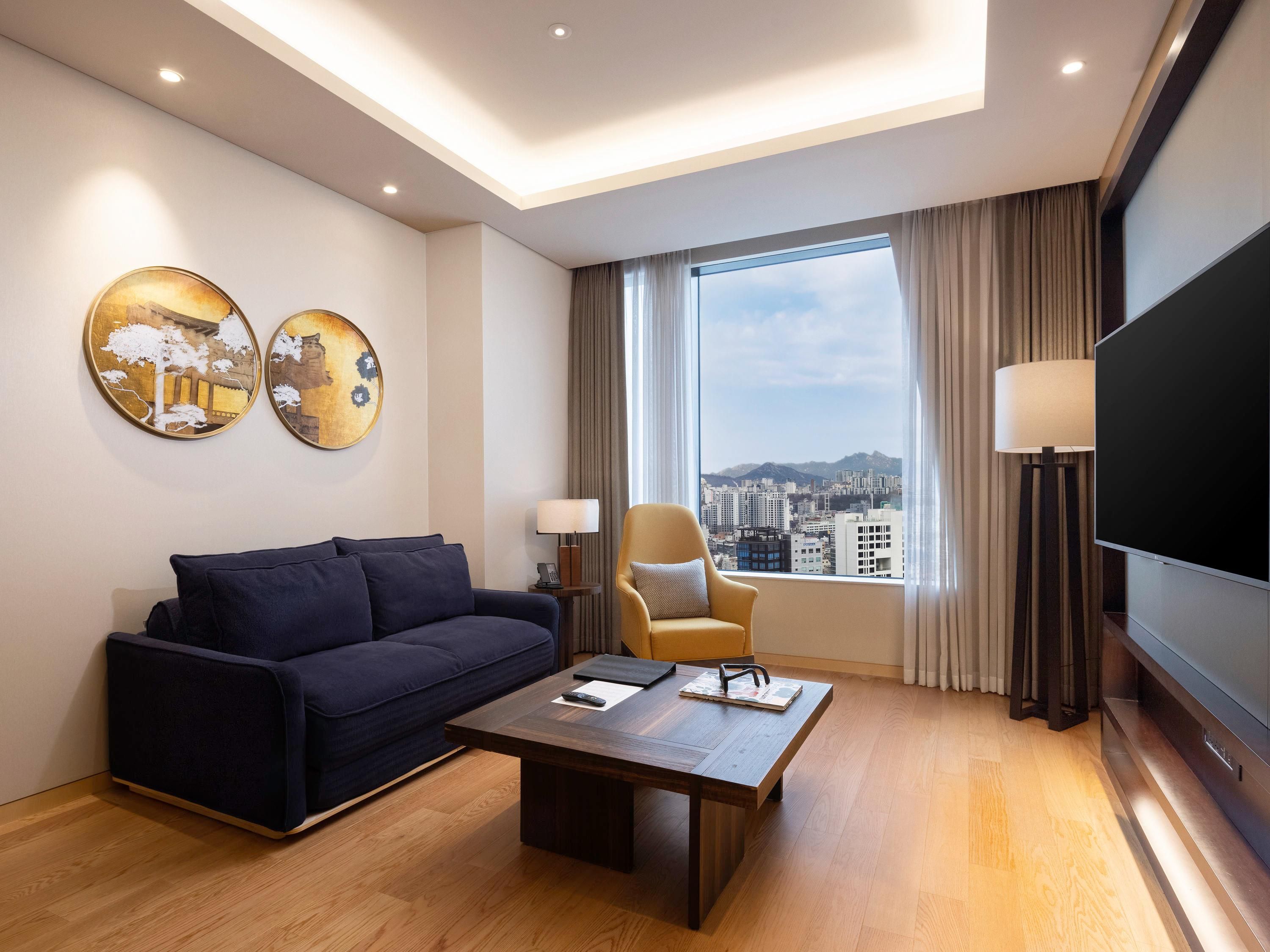 A modern hotel room with a sofa, a coffee table, a TV, and a large window with a city view.