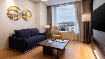 A modern hotel room with a sofa, a coffee table, a TV, and a large window with a city view.