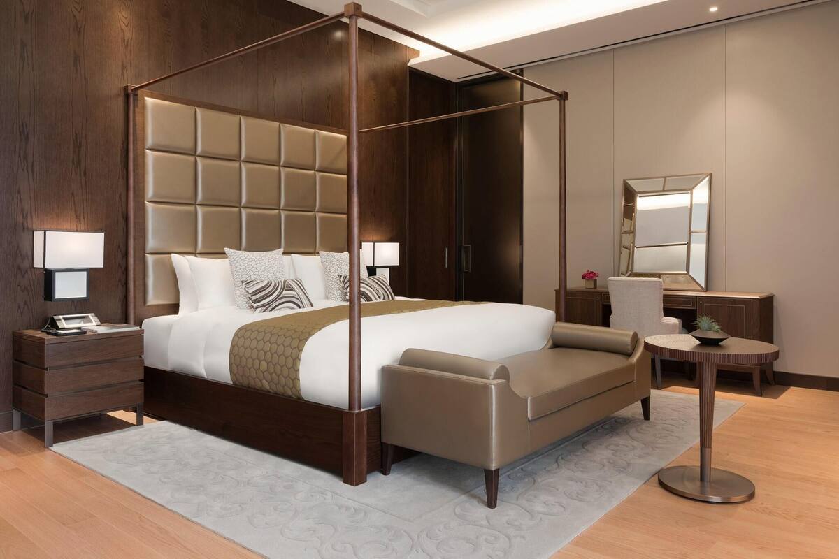 A four-poster bed with a tufted headboard, bedside tables with lamps, a round side table, and a mirror on the wall.