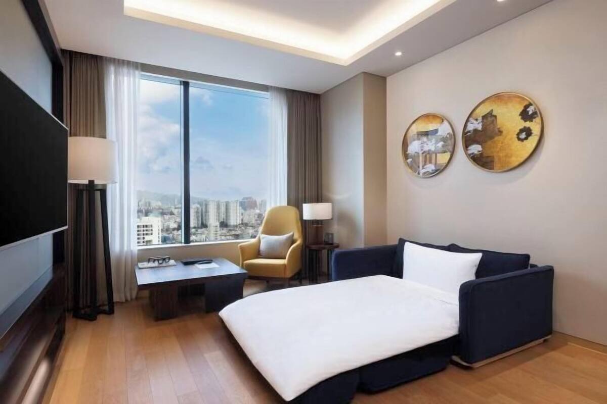 A hotel room with a bed, a sofa, a chair, a TV, a desk, and a large window with a city view.