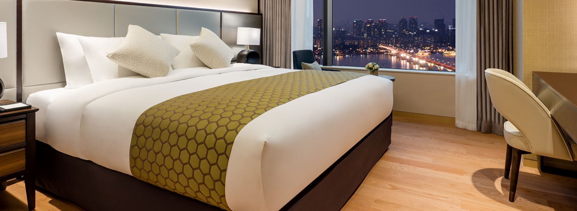 Une chambre d’hôtel avec un grand lit, un bureau, une chaise et une vue sur le paysage urbain illuminé la nuit.