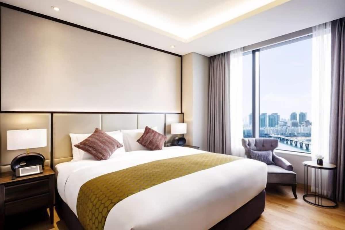 A hotel room with a large bed, a city view, a chair, and a small table.