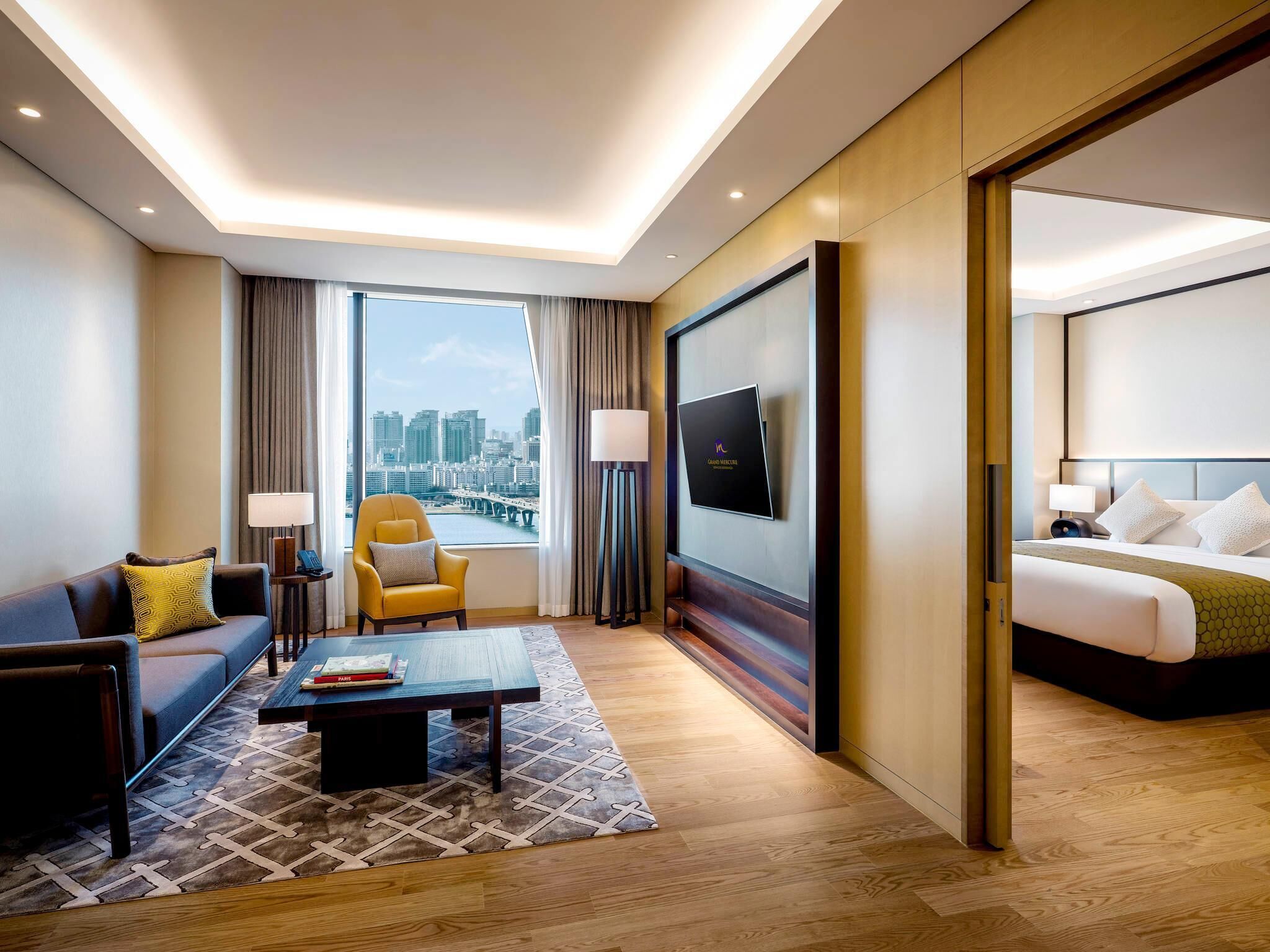 Une chambre d’hôtel comprenant un lit, un canapé, une chaise, une table basse, une télévision et une grande fenêtre donnant sur la ville.
