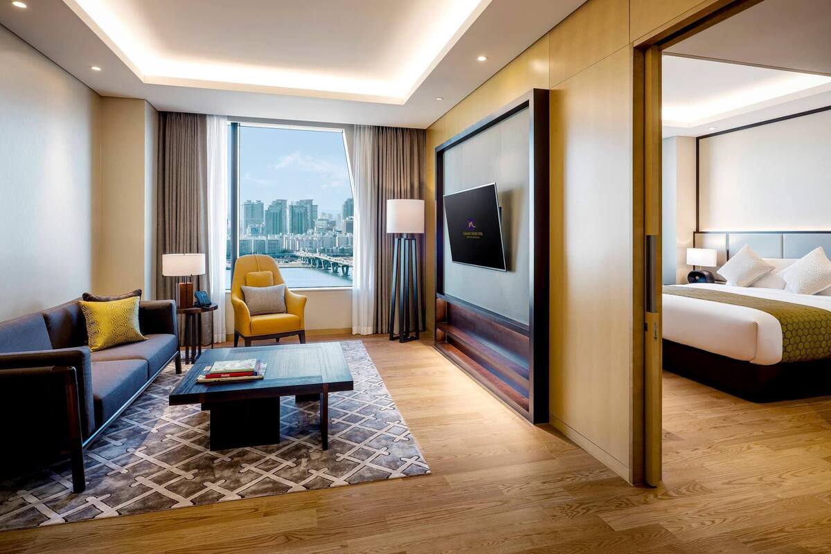 A hotel room with a bed, a sofa, a chair, a coffee table, a TV, and a large window with a city view.