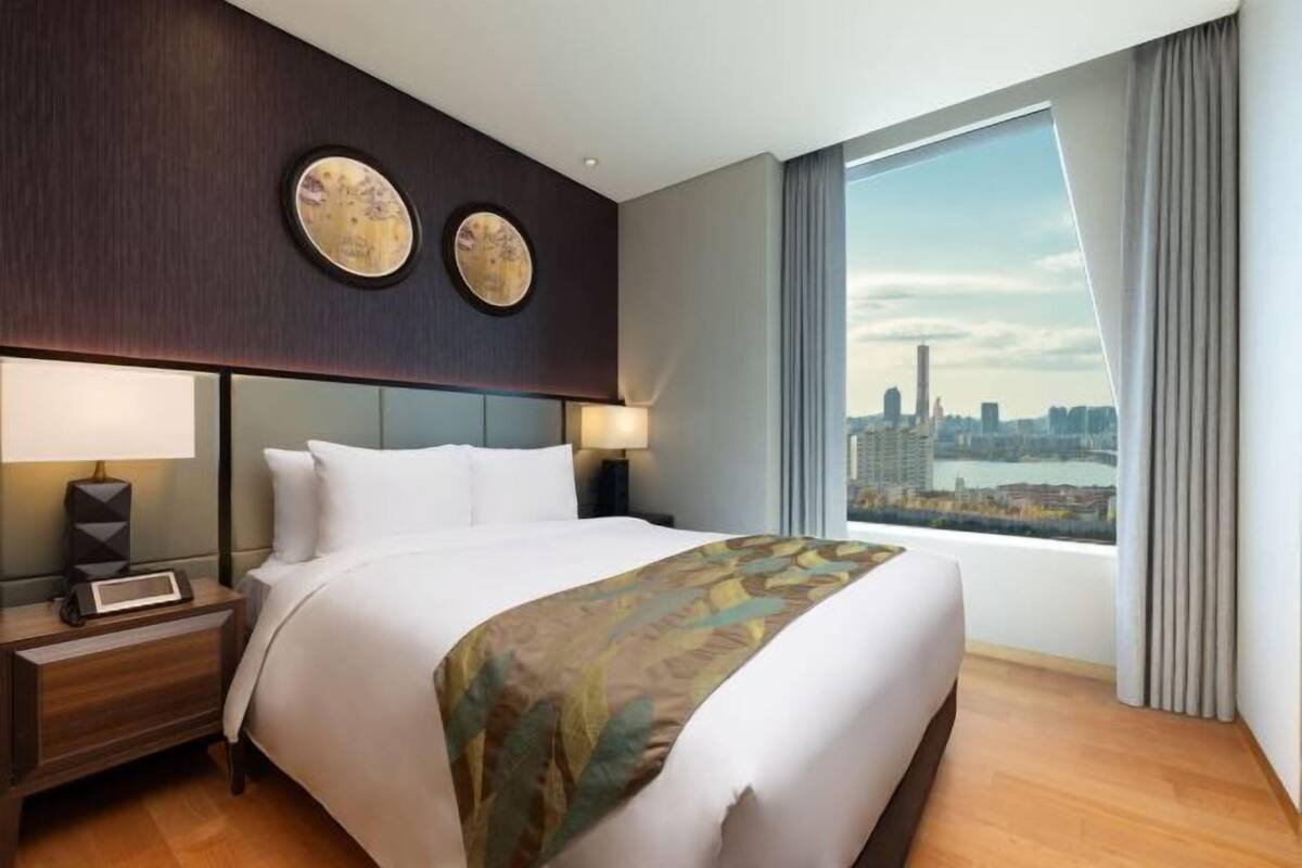 A hotel room with a large bed, a nightstand, a lamp, two wall art pieces, and a view of a cityscape through a large window.