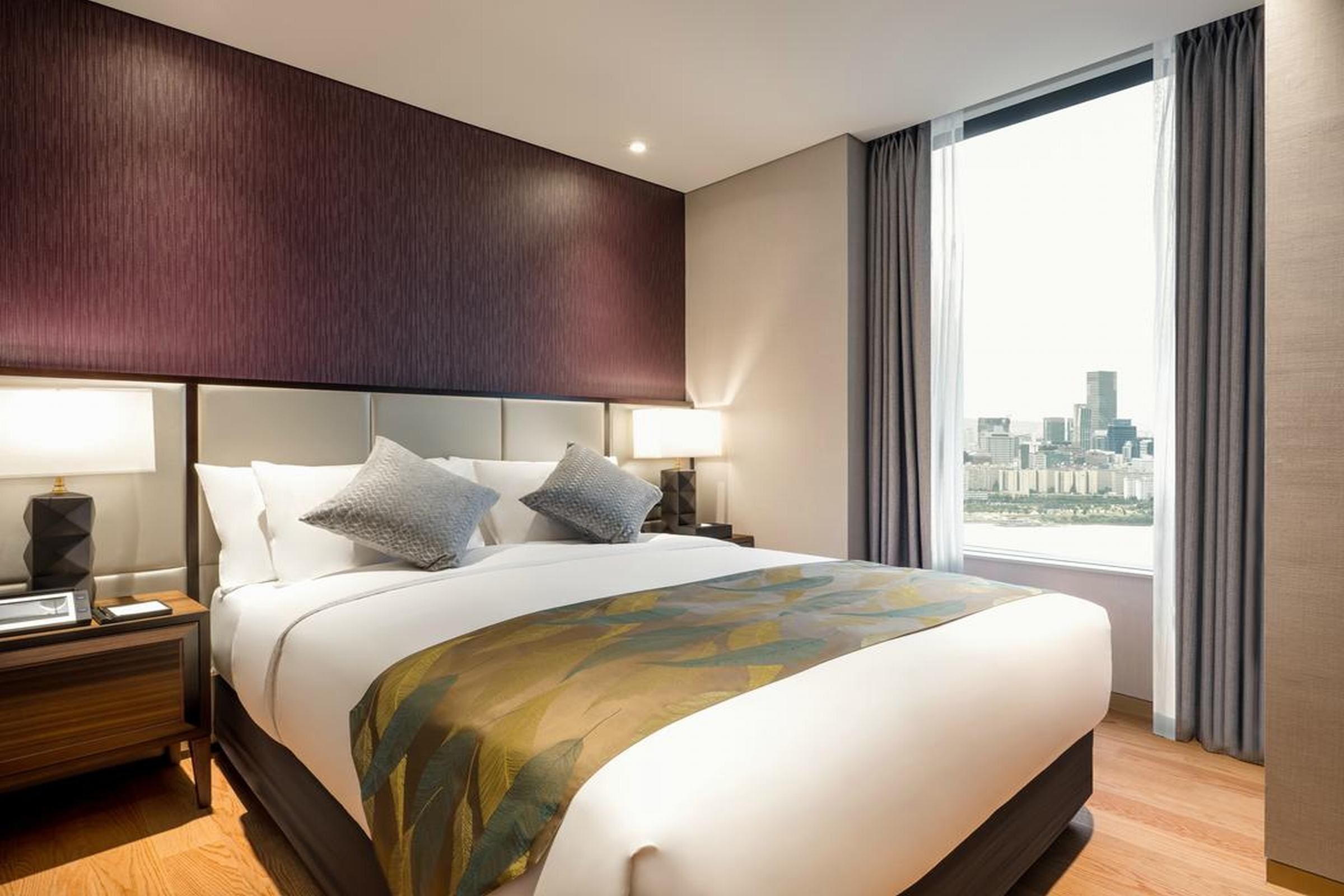 A hotel room with a large bed, a nightstand, a lamp, and a view of a cityscape through a window.