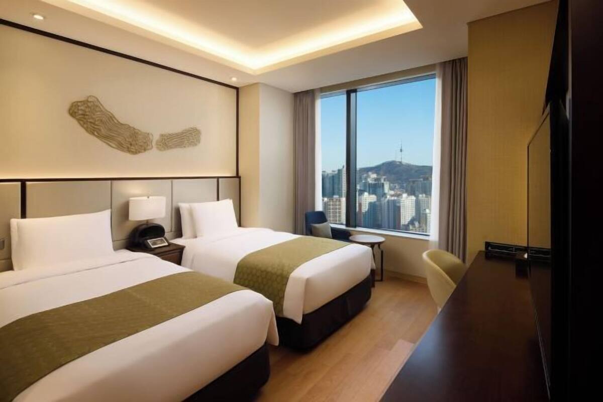 A hotel room with two beds, a desk, a chair, a TV, and a large window with a city view.
