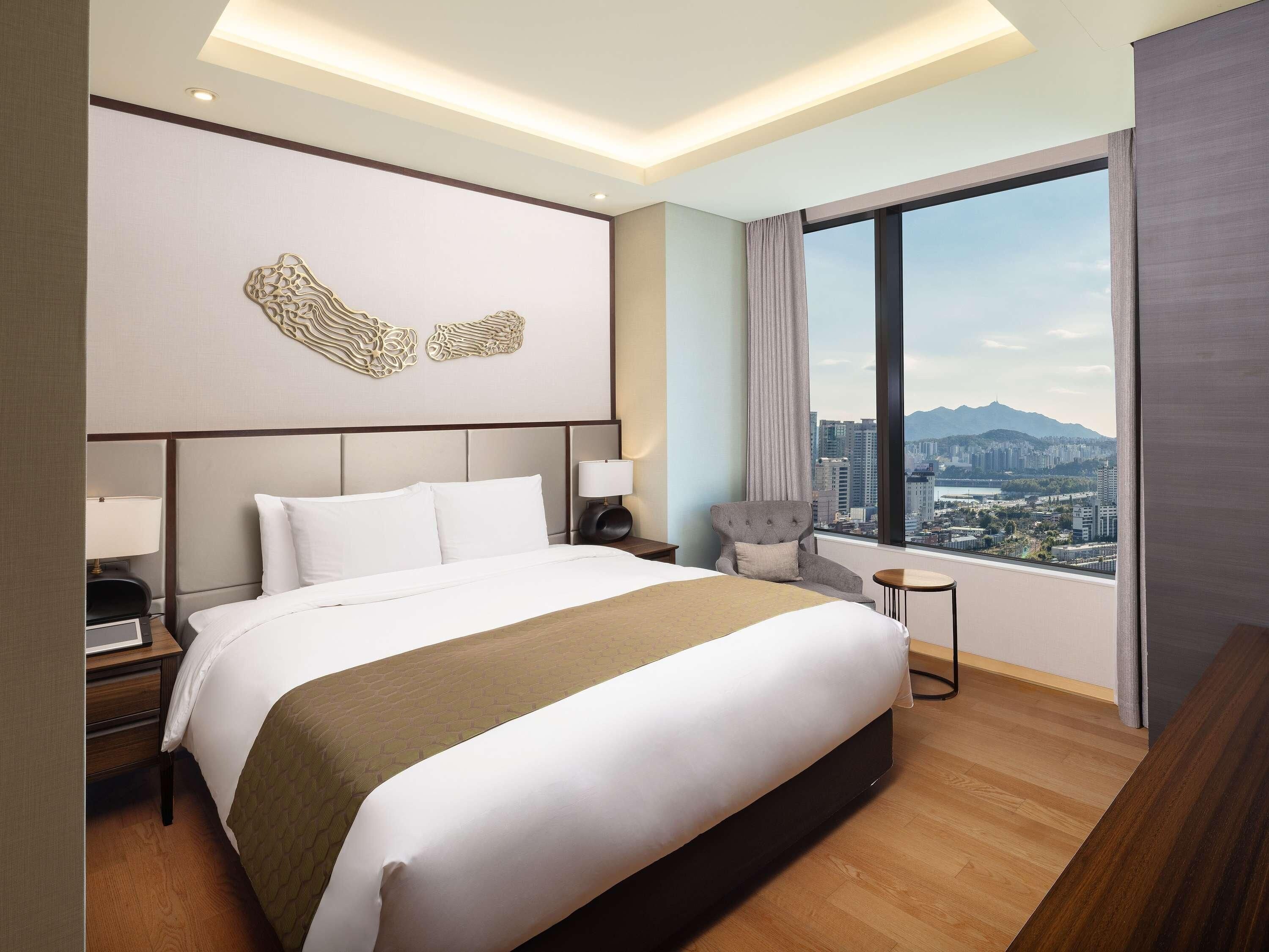 A hotel room with a large bed, a chair, a small table, and a view of a cityscape through a large window.