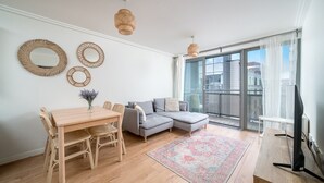 En moderne stue med et træspisebord, en grå sofa og et stort vindue med udsigt til bygninger.