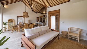 Uma sala de estar ampla com teto de madeira, um sofá, móveis de vime e uma cozinha ao fundo.